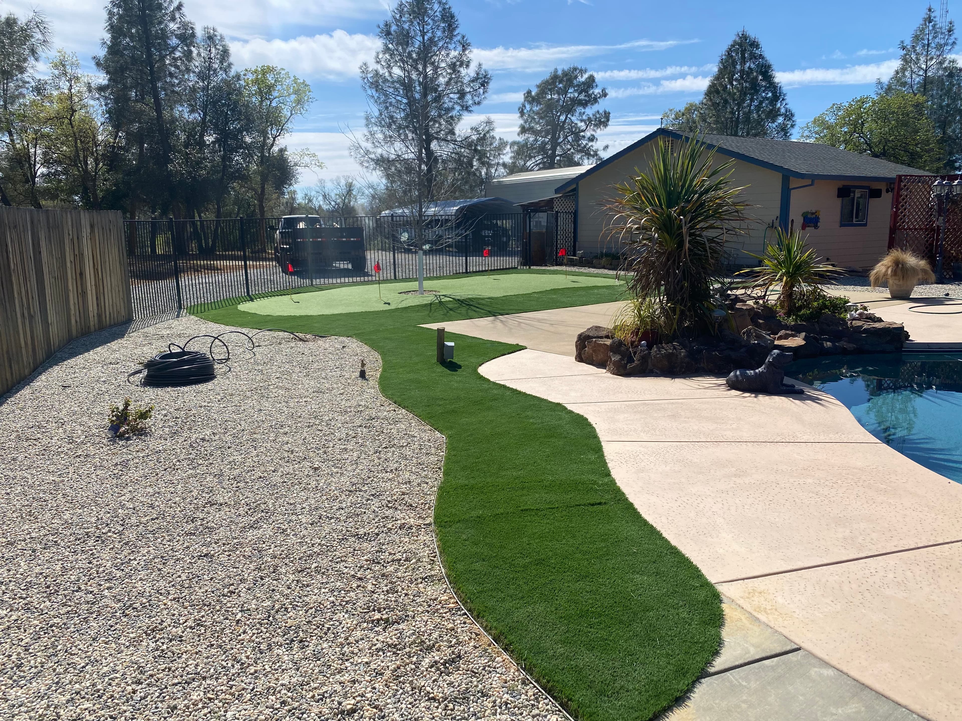Backyard featuring a synthetic putting green, gravel landscaping, and a swimming pool under blue skies.